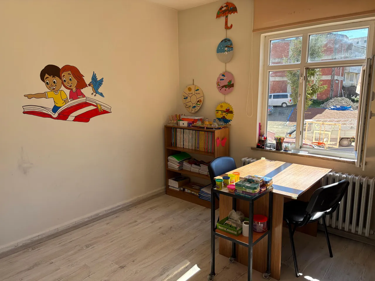 Aydınlık bir eğitim odası; duvarda uçan kitap üzerinde çocuklar resmi, masa ve kitaplık